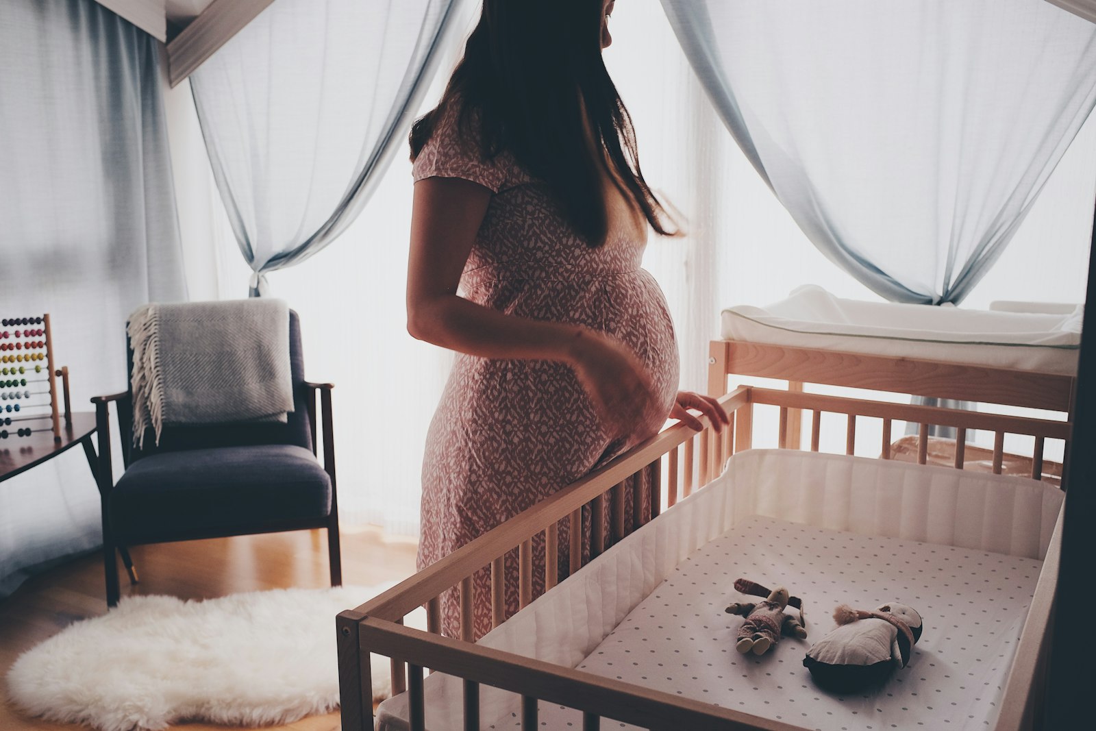 An expecting parent standing beside a wooden crib in a sunlit nursery